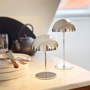 Two chrome-plated table lamps stand and mirror the room on a marble table; around them are stacked plates, cutlery and a black teapot in a bright kitchen with a slanted window.
