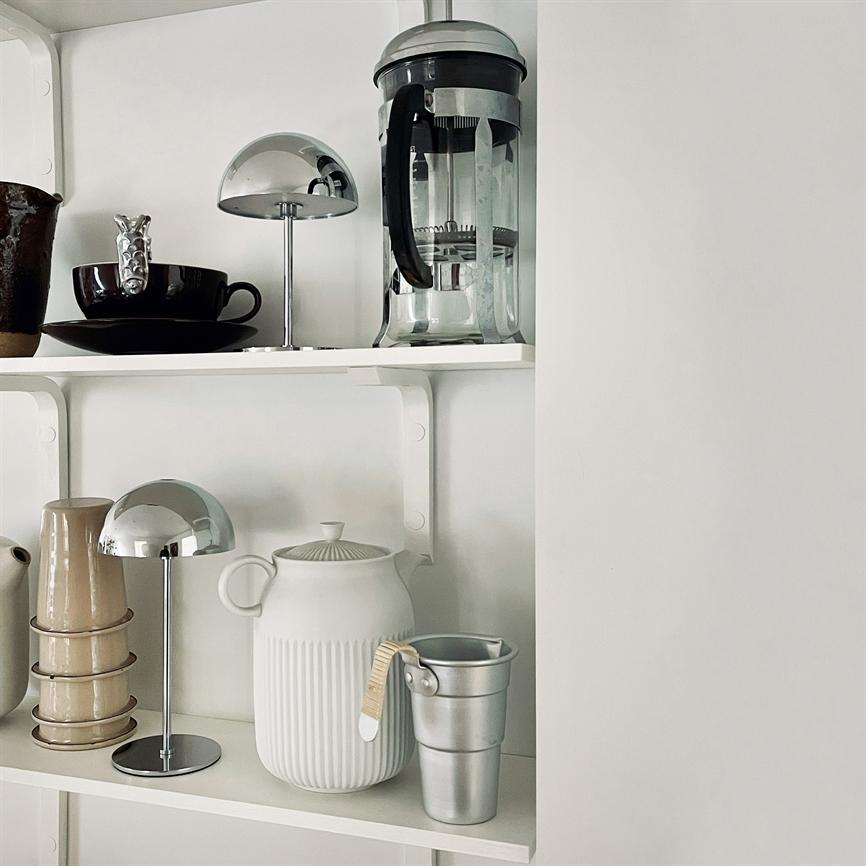 A white kitchen shelf, filled with kitchen utensils: a French press, a grooved white teapot, stackable beige mugs, small chrome lamps and a small aluminum tub — arranged calmly against a white wall.