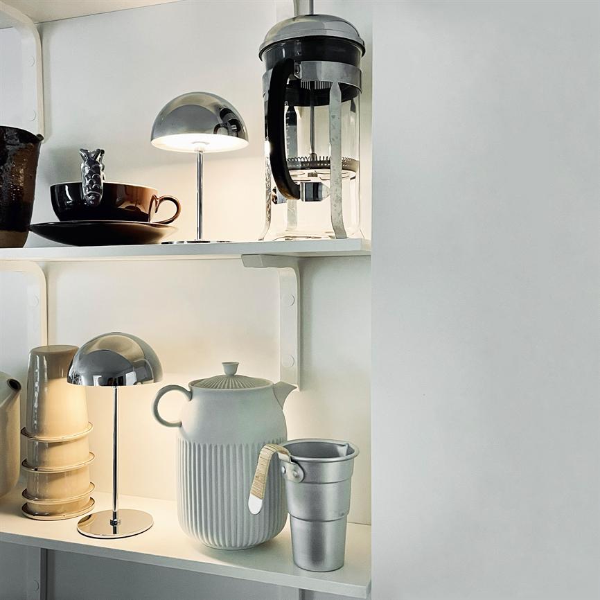 Ceramic ribbed teapot stands in the middle of a white shelf; surrounded by a chrome table lamp, a small metal goblet, stacked mugs and a French press in a bright kitchen shelf environment.