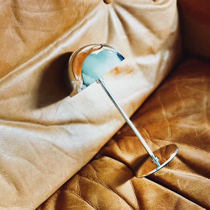 A chrome table lamp lies to the side, slightly tilted and reflecting the surroundings; its slender steel shaft and hemispherical shade rest on wrinkled, golden-brown leather on a couch.