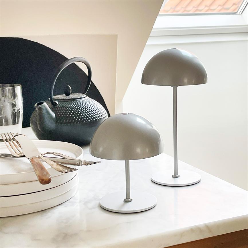 Two grey, mushroom-shaped table lamps stand on a marble table; in the background a black cast iron teapot, a stack of white plates with cutlery and a glass by a slanted window.