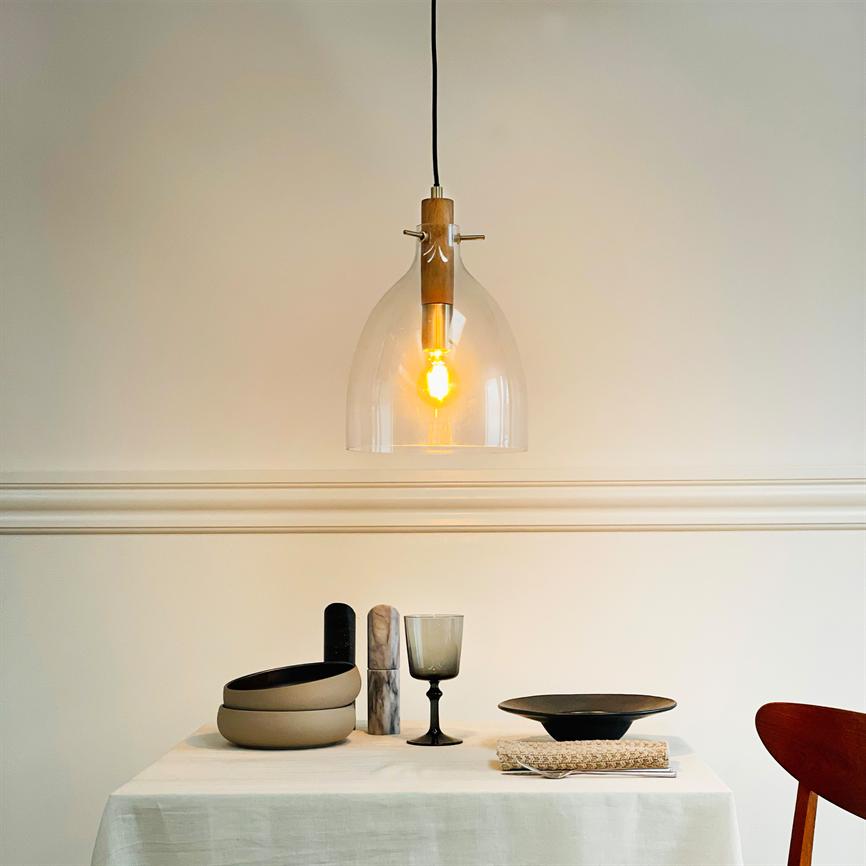 A glass pendant light radiates warm light over a white-clothed table; the table holds neutral bowls, a dark wine glass, a flat bowl and a wooden chair in a bright, minimalist dining room.