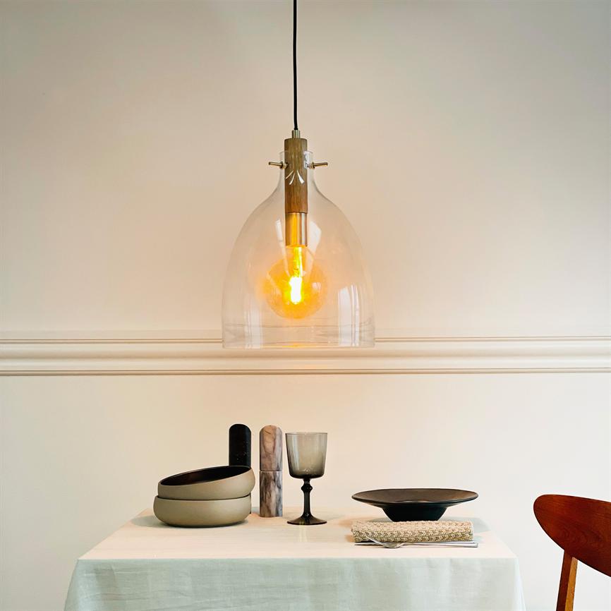A glass pendant lamp hangs and emits a warm orange glow above a simple set table with bowls, a wine glass, a black plate and napkin in a bright, minimalist room.