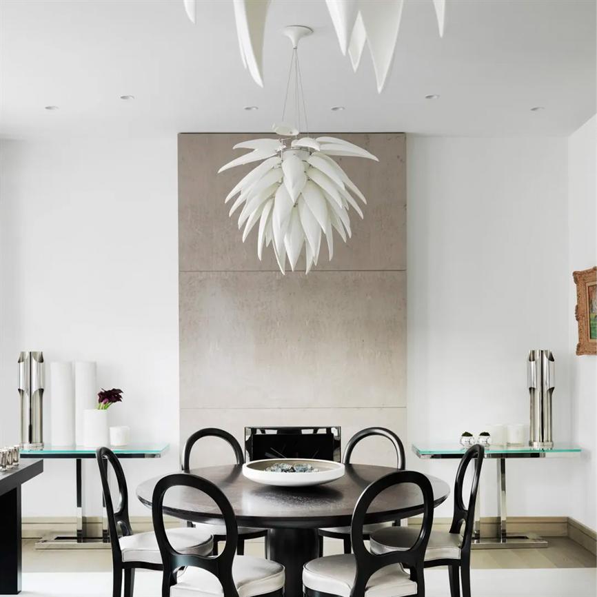 Chandelier in white, leaf-like shades hang above a round dark dining table; a bowl in the middle of the table, surrounded by black chairs in a bright, minimalist dining room with glass tables.