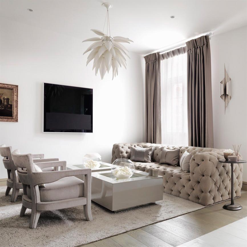 Large button-upholstered corner sofa dominates the room, stands by the window; two matching armchairs and low coffee table with glass balls in front, wall-mounted TV, long curtains and decorative hanging lamp in light neutral tones.