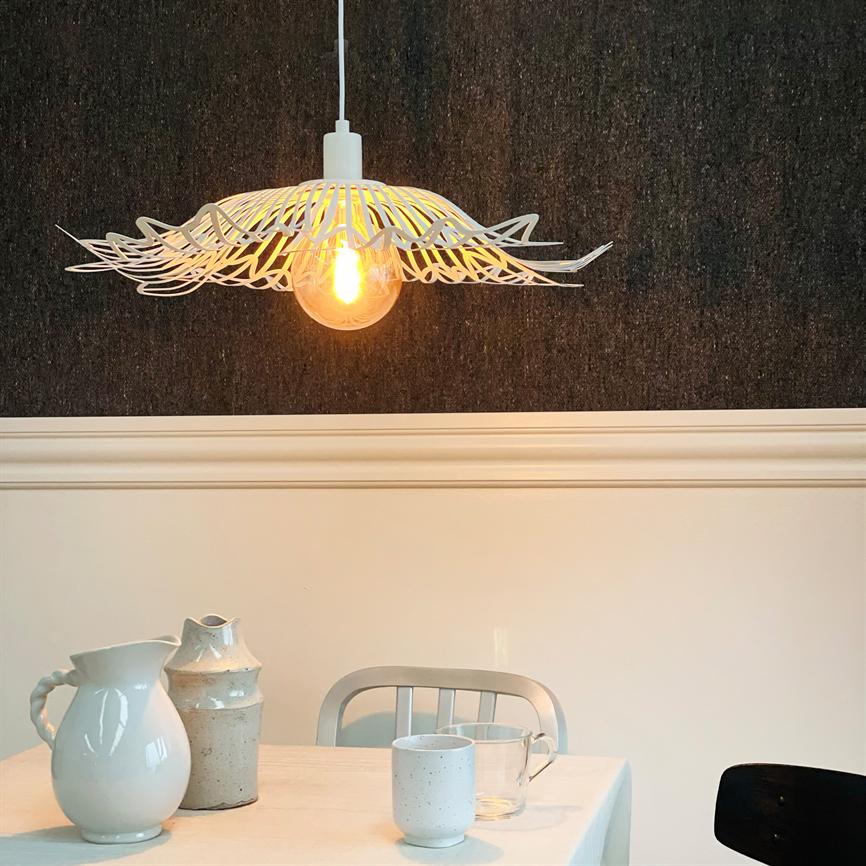 A decorative white pendant light hangs lit above a dining table; its filament bulb casts warm light, while a table with bright jugs, cups and a metal chair stands against a dark wallpaper wall.