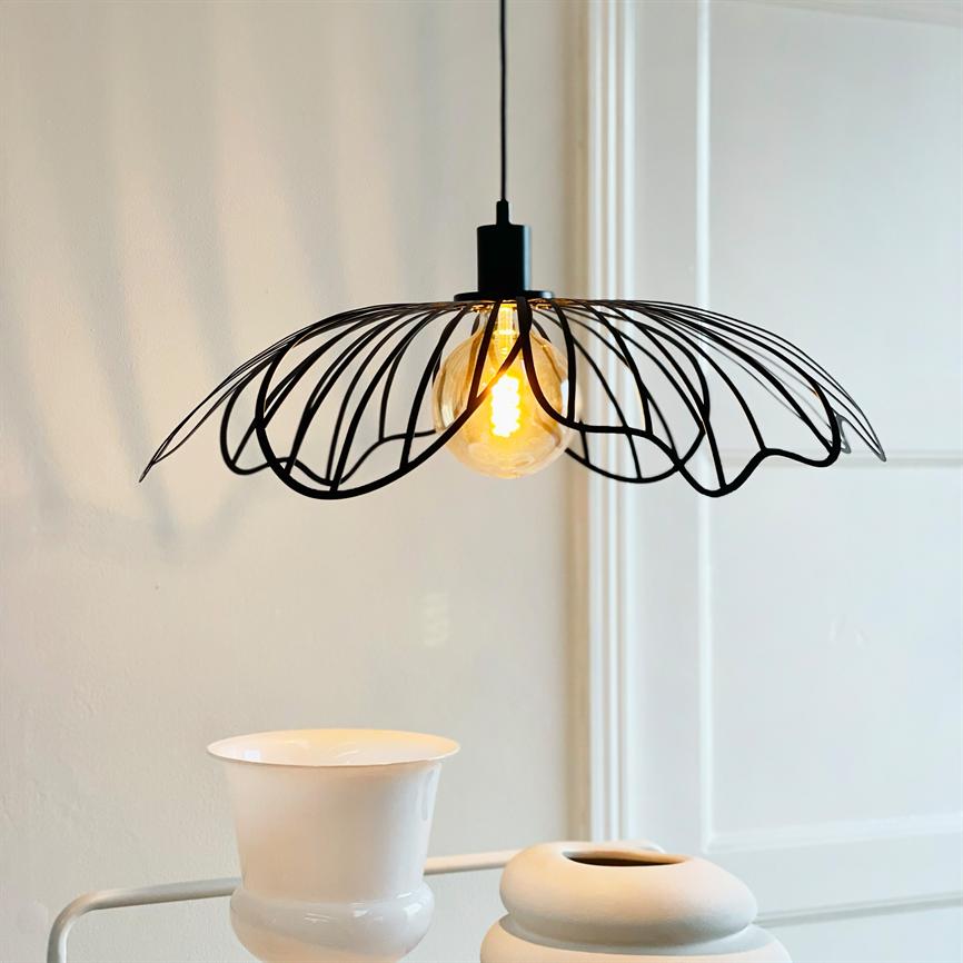 A black, leaf-inspired ceiling lamp hangs and casts a warm glow from a visible light bulb over two white vases on a table in front of a light wall and a door.