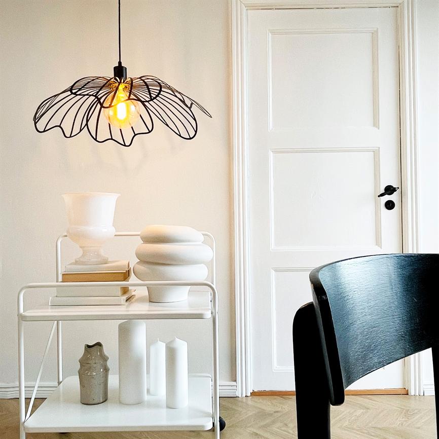 A black, wire-shaped ceiling lamp casts warm, golden light over a white cart with ceramic vases in front of a white door and a black wooden chair in a bright, minimalist room.