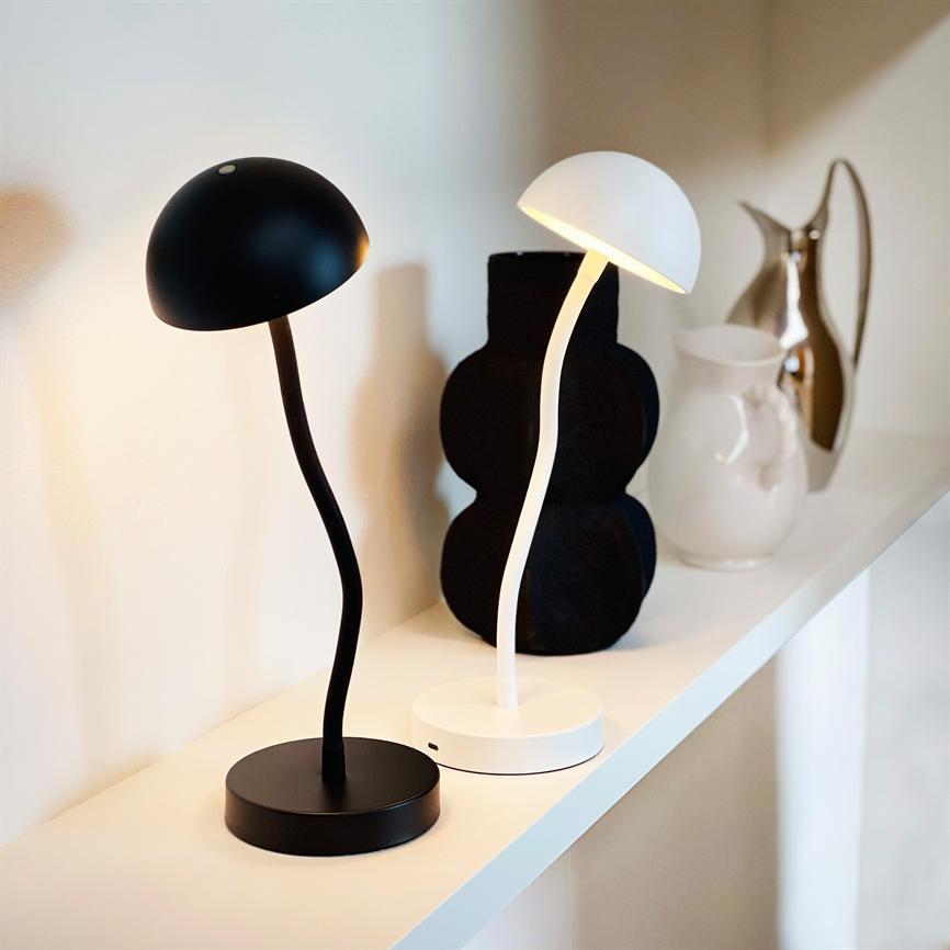 Two mushroom-shaped table lamps — one white, dimly lit, and one black, turned off — stand on a white shelf in front of a neutral wall with decorative vases.