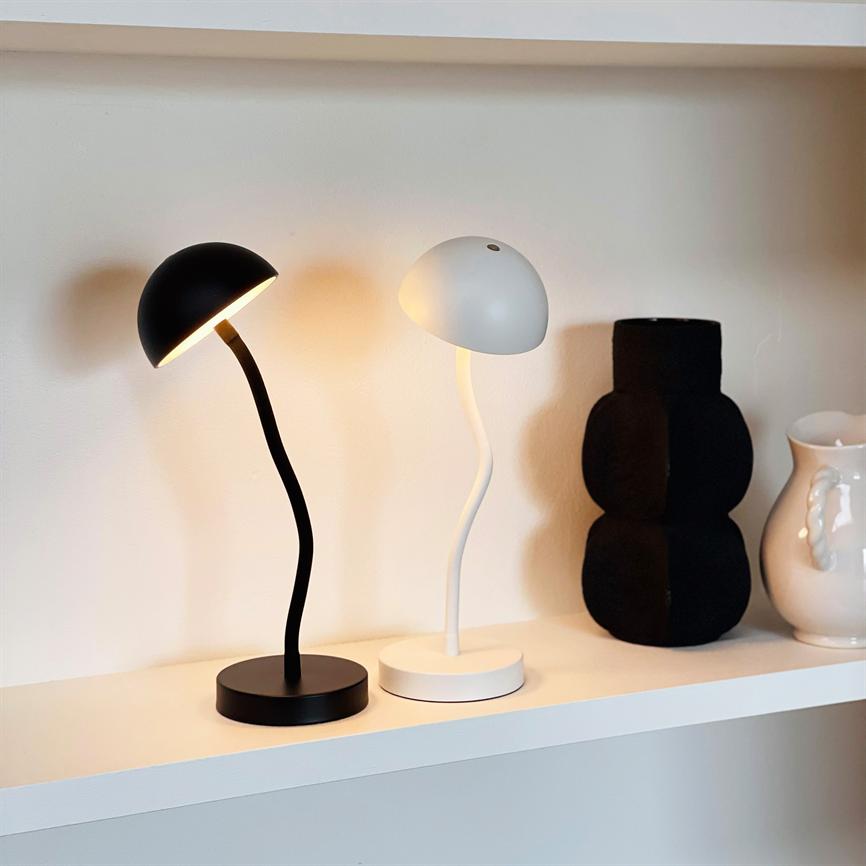 Two curved table lamps—one black, lit, and one white, off—stand on a white wall shelf in front of a cream-colored wall, flanked by a black sculptural vase and a white pitcher.