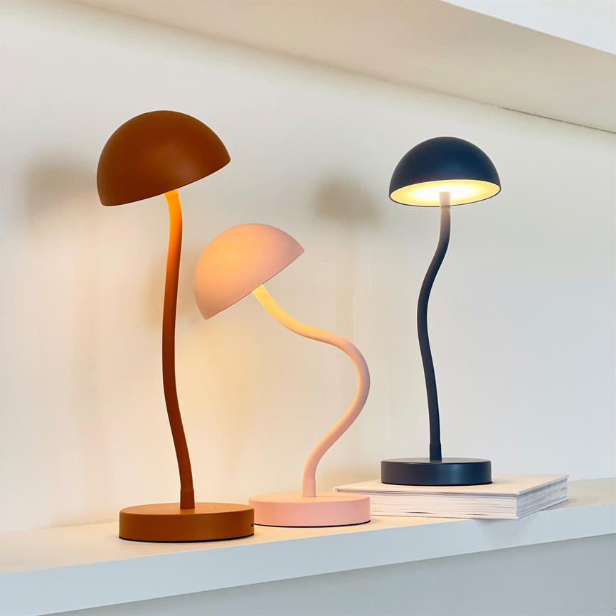 Three decorative table lamps stand on a white shelf and glow softly; each has a curved shaft and shade in different colors, one stands on a stack of books.