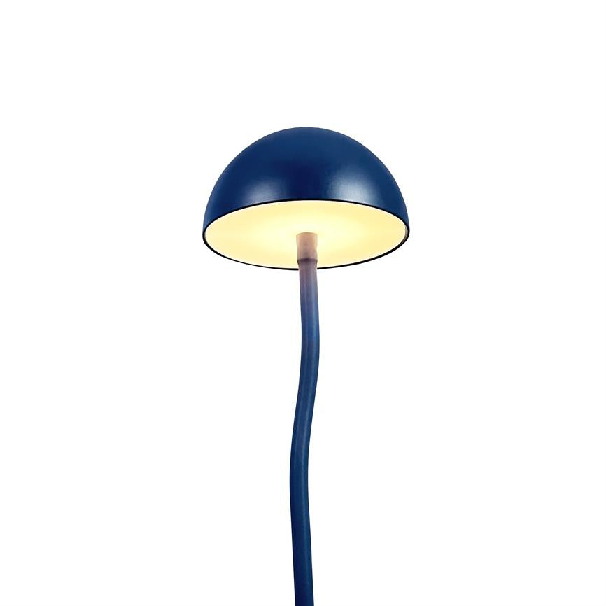 Blue table lamp displays a hemispherical shade, lit from within; a slim, slightly curved bar leads downwards against a neutral white background.