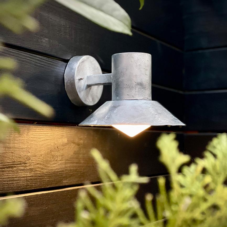 Wall lamp lights downwards; gray metal fixture mounted on dark, horizontal wooden boards with blurred green plant life in the foreground, warm light casting a glow on the surface of the wood.