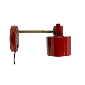 Red wall lamp extends a brass arm with cylindrical shade to the right on a pure white background; black power cable hangs down from the wall bracket.