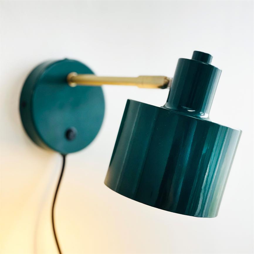 Dark green metal wall lamp hangs angled downwards on a brass arm; round wall bracket and black cord are seen against a light, white wall.