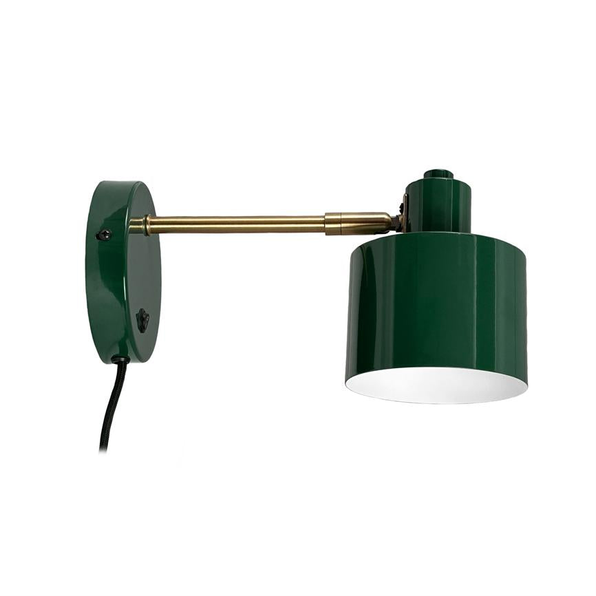 Green metal wall lamp extends a brass arm with a downward cylindrical shade, mounted on a round wall plate against a white background.
