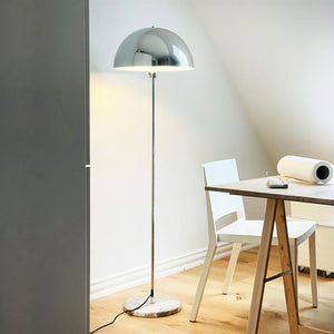 Floor lamp with shiny chrome dome stands lit on a marble base and illuminates a corner with white walls; next to it is a white chair and a wooden table with a roll of paper and a telephone.