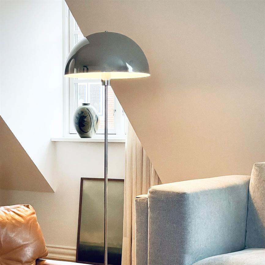 Floor lamp glows softly; standing in front of a slanted window with a ceramic vase on the sill; surrounded by a light gray sofa and a brown leather corner in a quiet, modern living room corner.