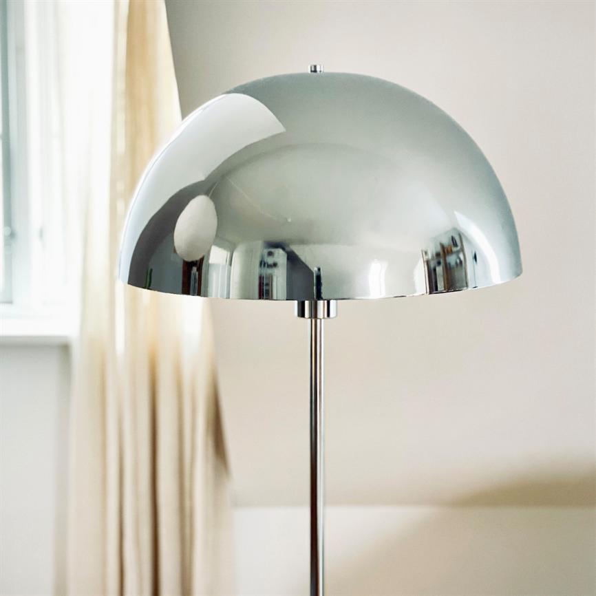 A chrome hemisphere lamp reflects a bright space; it stands on a slender pole by a light-curtained window in a neutral, calm interior.