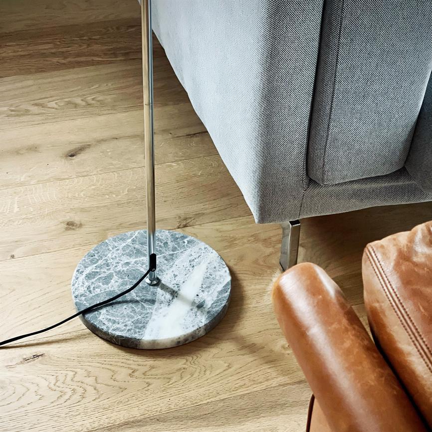 A floor lamp with a round marble base stands next to a gray upholstered sofa; chrome rod and black wire lie on a light wooden floor, close to a brown leather chair.