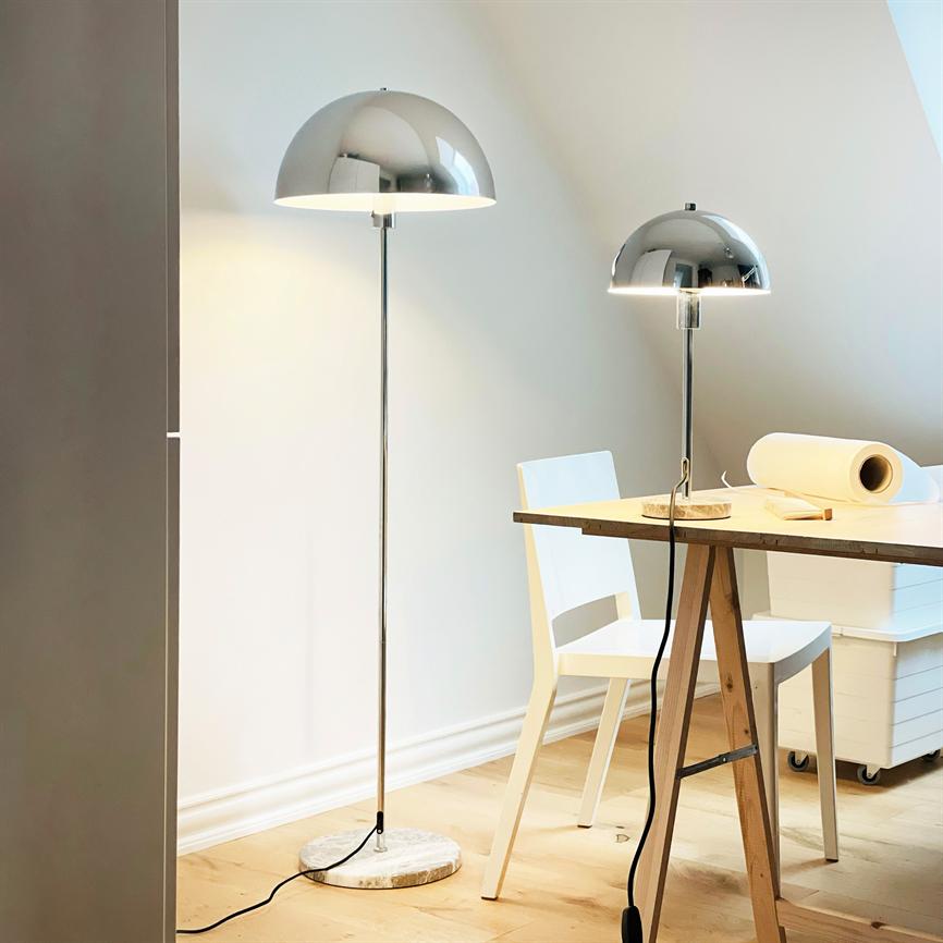 Two chrome dome lamps — a floor lamp and a table lamp — shine softly above a wooden table; by the table is a white chair in a bright, minimalist room with a wooden floor.