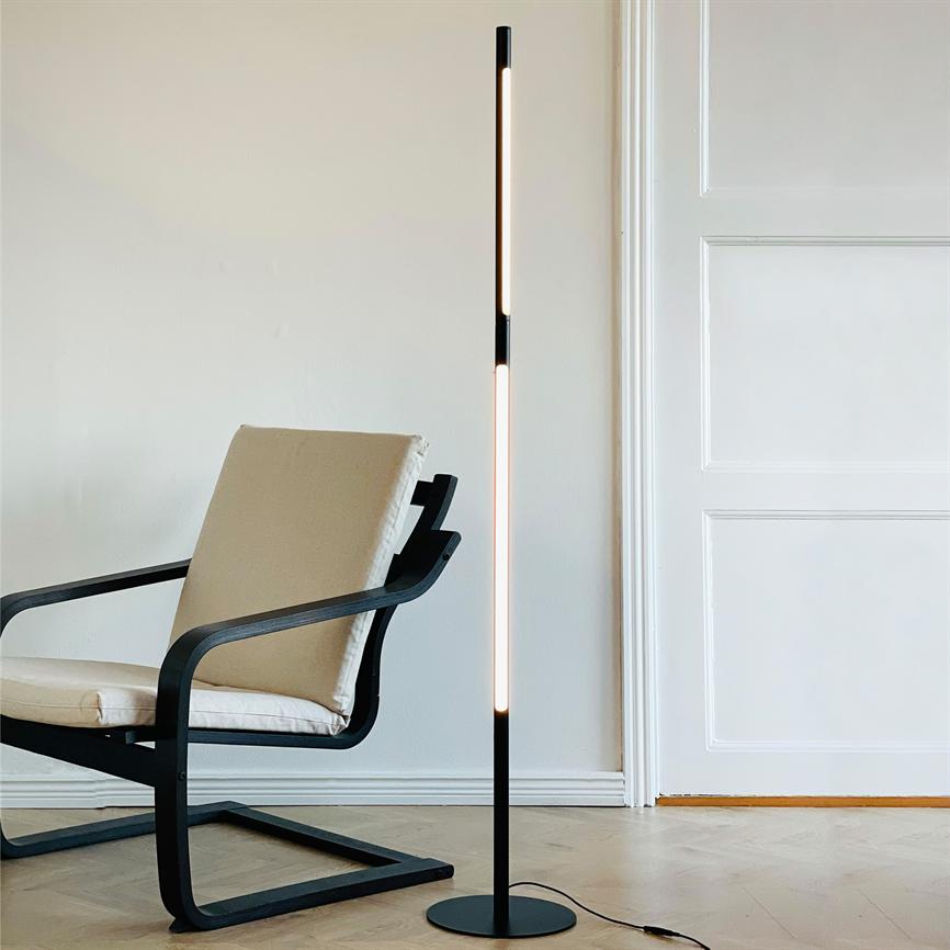 Slim black floor lamp emits soft, warm light next to a beige upholstered armchair in a bright room with white walls and wooden floor in front of a white door.