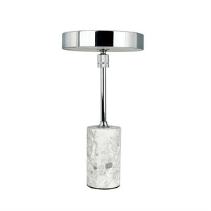 Table lamp stands vertically with round, shiny chrome shade and slim rod; it rests on a cylindrical white-gray marble base against a uniform white background.