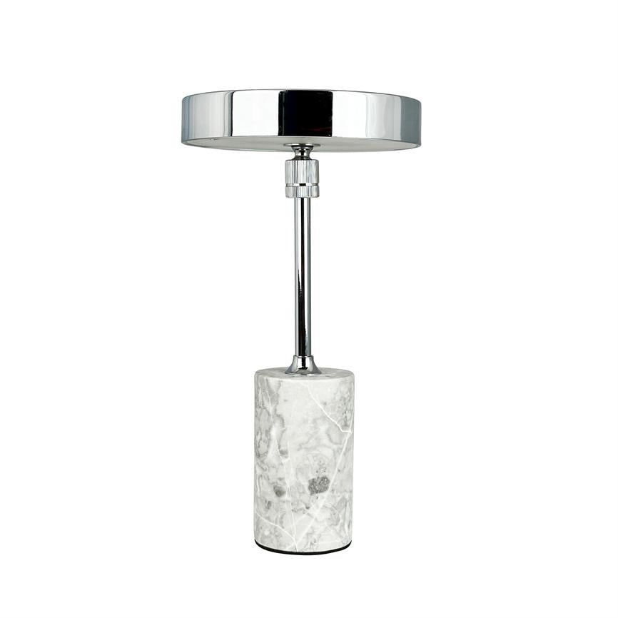 Table lamp stands vertically with round, shiny chrome shade and slim rod; it rests on a cylindrical white-gray marble base against a uniform white background.