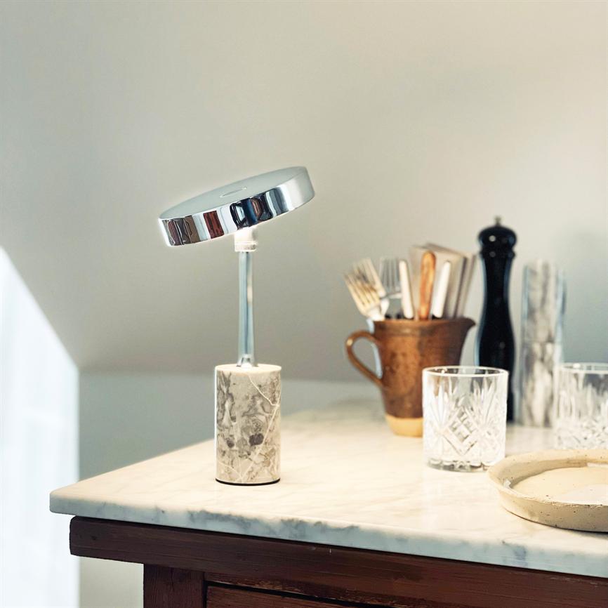 A modern table lamp with a round chrome shade and marble base stands on a marble table top; in the background mug with cutlery, glass, pepper grinder and a small dish.
