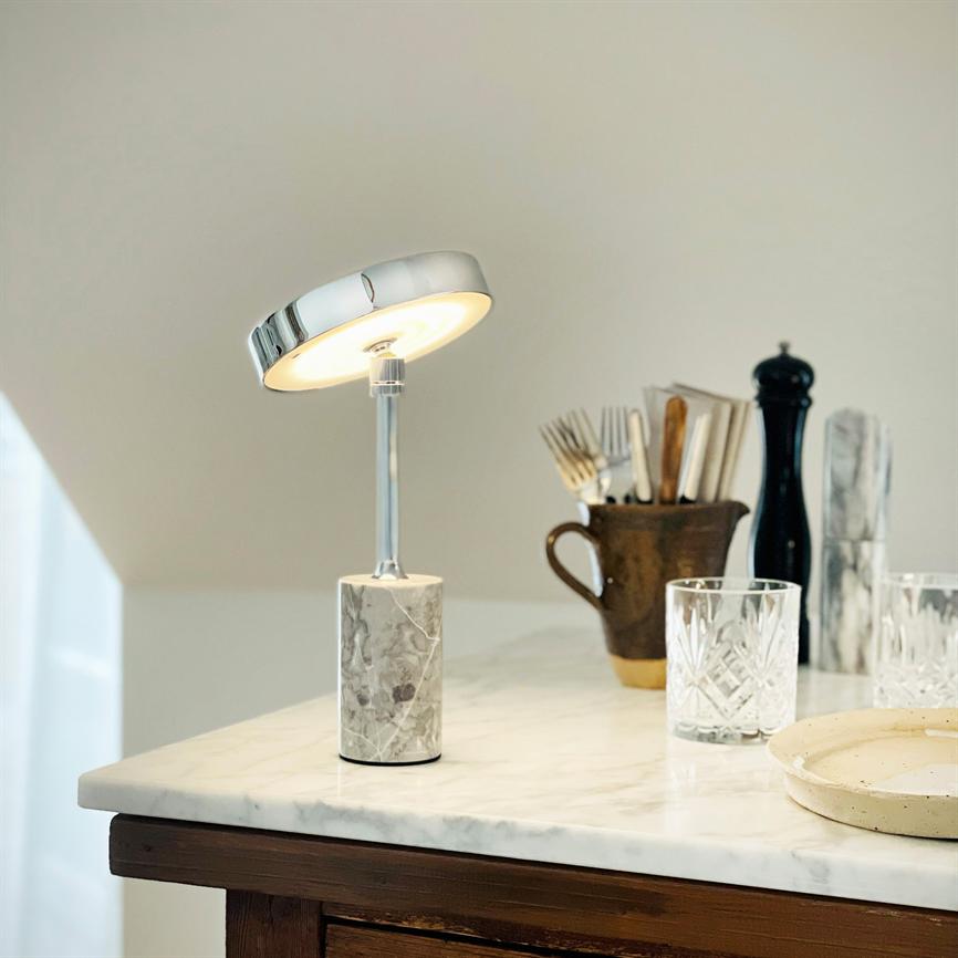 Table lamp with round chrome shade and marble base glows softly; standing on white marble tabletop in a kitchen/serving area with glasses, cutlery in a jar and pepper grinder in the background.