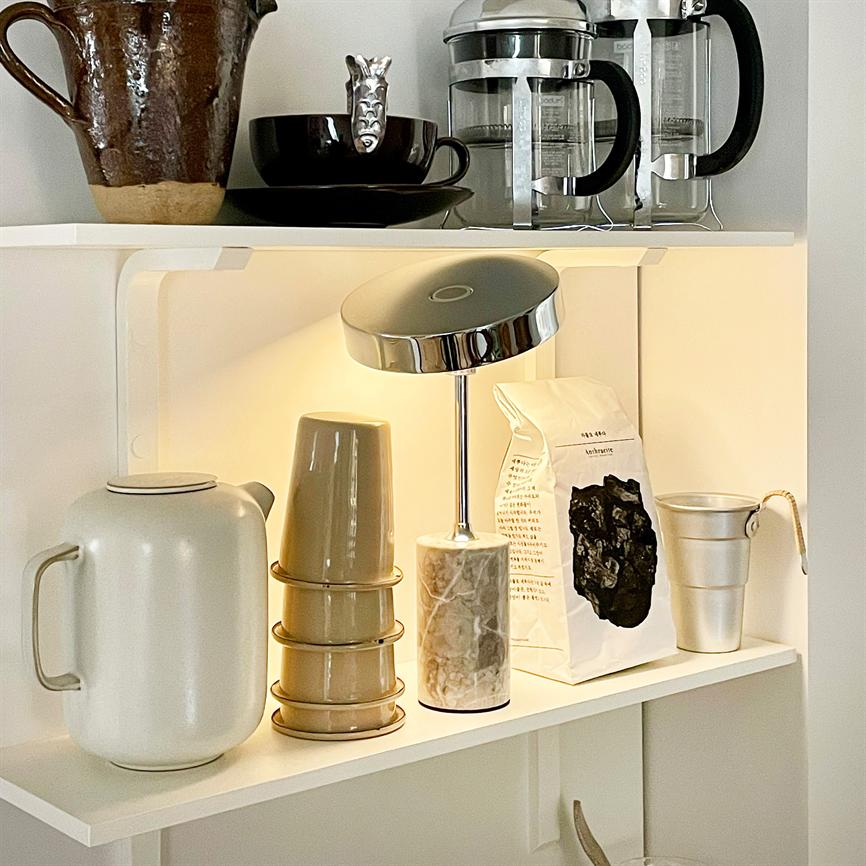 A small table lamp shines on a white shelf, surrounded by a ceramic jug, stack of cups, a coffee bag, metal cup and two French press jugs in a bright kitchen corner. No readable text.