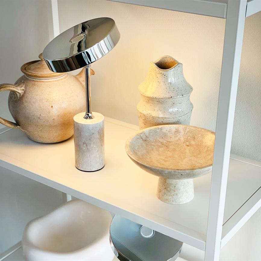 Chrome lamp is lit and casts warm light on a white shelf; around it are dusty beige ceramic pots and a standing bowl in a neutral, minimalist interior.
