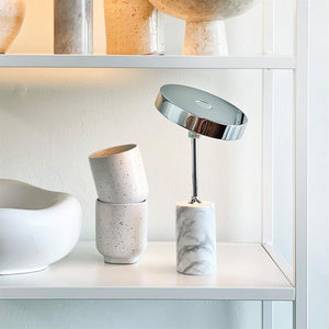 Table lamp with round chrome shade tilts on a slim rod above a marble cylindrical base, placed on a white shelf next to two stackable ceramic mugs and a bowl.