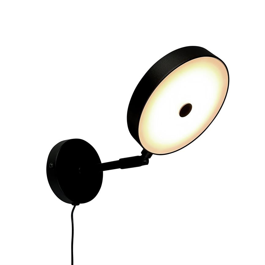 Wall lamp with round, illuminated shade, shines with warm, diffused light; black arm and wall base are mounted on white background, arm can be adjusted, black cord hangs down.
