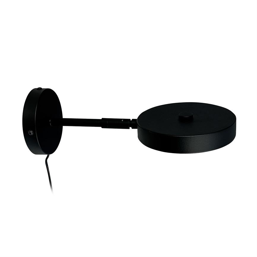 Black wall lamp with round wall base; a straight arm leads to a flat, circular lamp head, cord hanging down, against neutral white background.