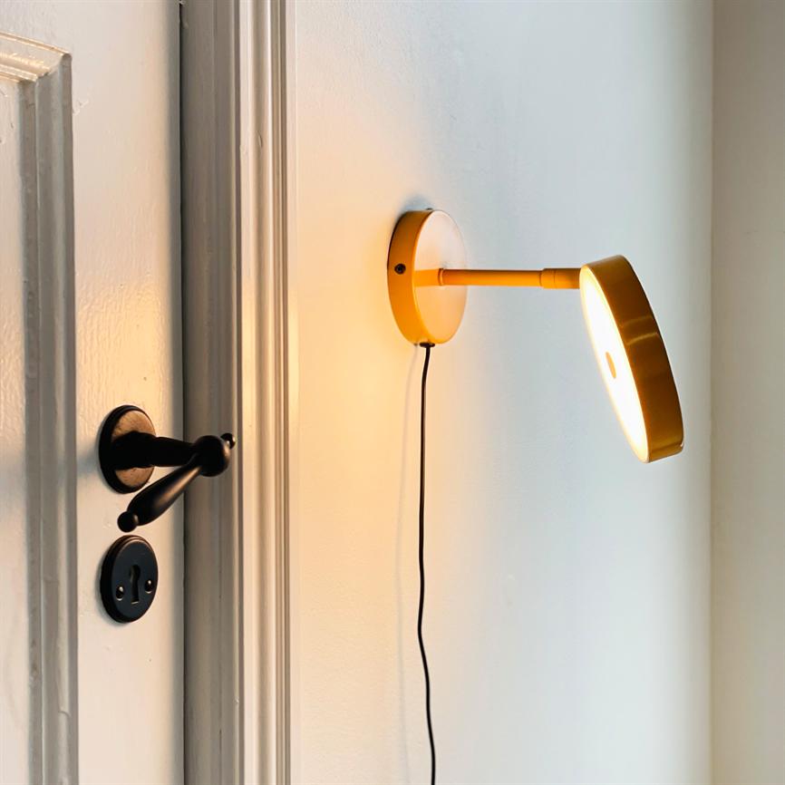 Yellow wall lamp emits warm light, mounted with wire downwards, illuminates a white wall close to a white door with a black door handle.