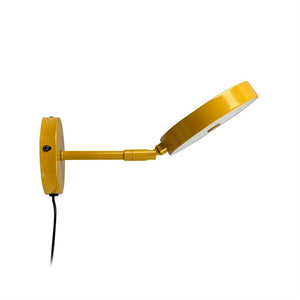 Yellow wall lamp mounted on round base, with extended arm and adjustable round lamp head angled downwards; black power cable hangs from the base against a white background.