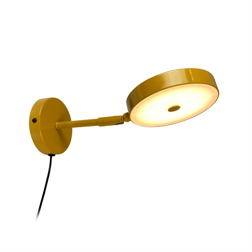 Wall lamp in yellow metal radiates warm light from a round LED disc, mounted on an outward arm from a round wall bracket with a visible black cord.