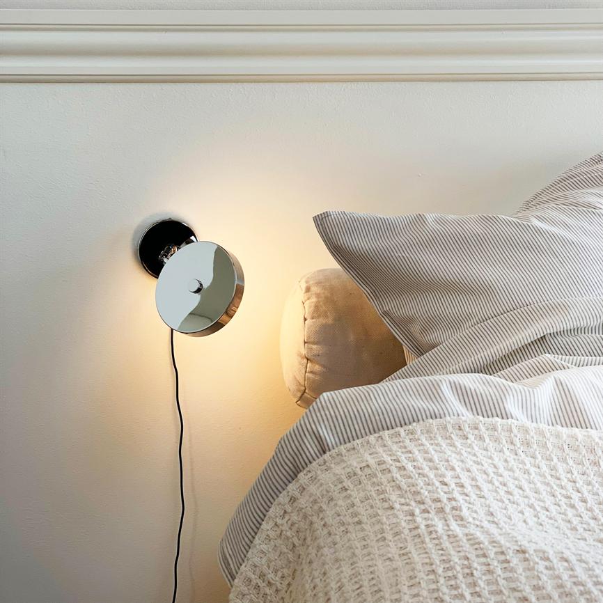 A wall lamp glows dimly above a bed; chrome round lamp with visible cord, beige pillow and striped sheet and a light, woven quilt against a neutral wall.