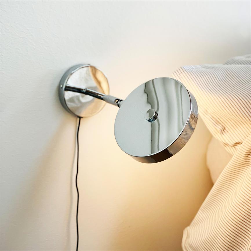 Hanging on a white wall, the chrome wall lamp shines warmly downwards and casts reflections on its shiny surface; it sits by a bed with bright, fine-striped pillows.