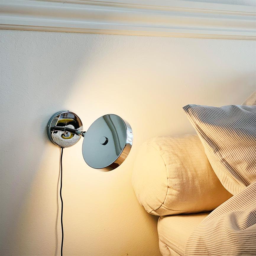 A chrome wall lamp casts warm, focused light; it is mounted above a beige sofa with round neck pillow and striped pillow in a bright, calm bedroom environment.