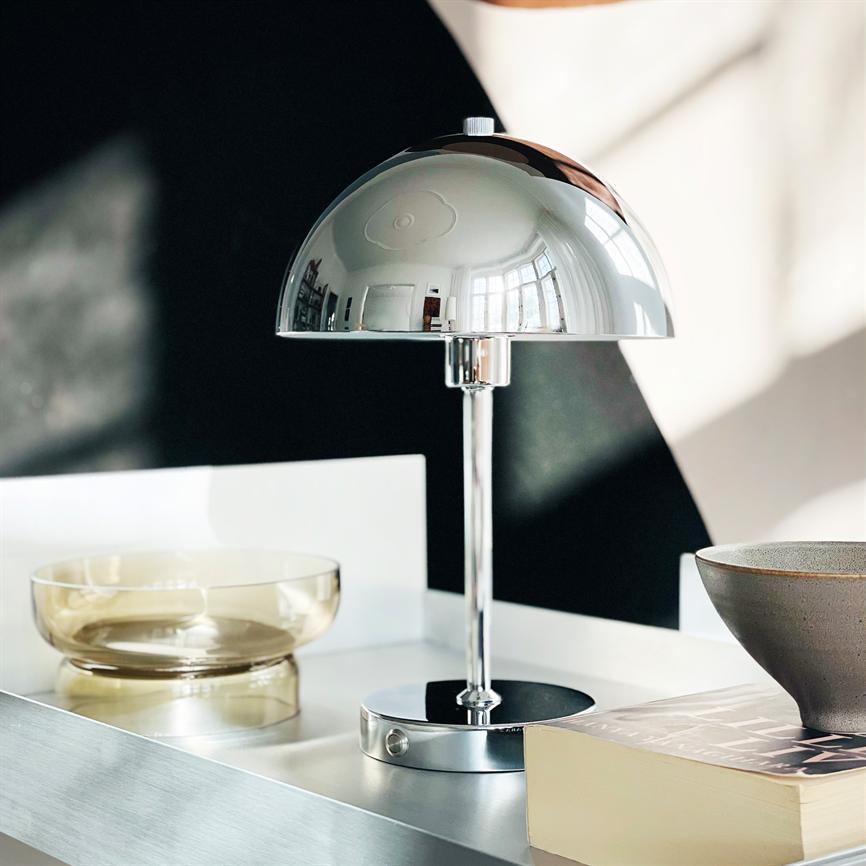 A chrome table lamp reflects the room, stands on a white tray next to a transparent bowl and a book; soft light and contrasting dark back wall in a bright, modern interior.