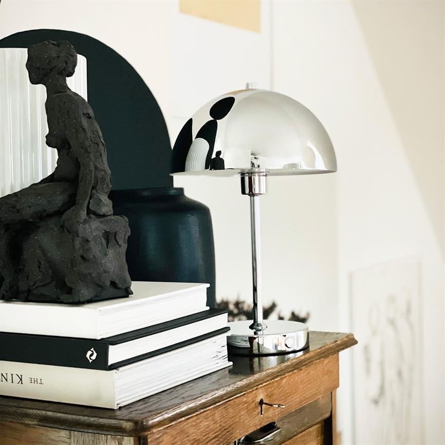 A chrome table lamp reflects the surroundings; it stands next to a dark sculpture on a stack of books atop a wooden cabinet in a bright, minimalist room.
 
 Text: "THE KIN"