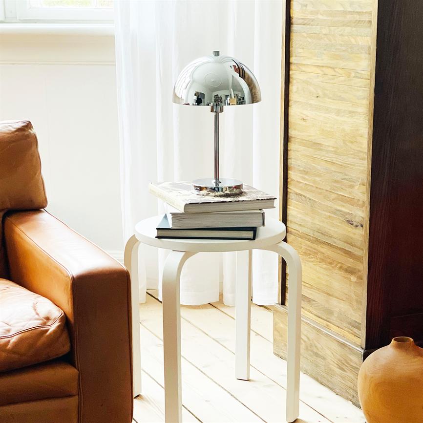 Chrome lamp stands on a stack of books on a small white side table; in a bright room by a cognac-coloured leather sofa, white curtain, light wooden floor and a wooden cupboard.
