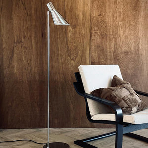 A chrome floor lamp stands next to an upholstered armchair with a cream cover and brown throw pillows, in front of a warm dark wooden wall and light patterned wooden floor.