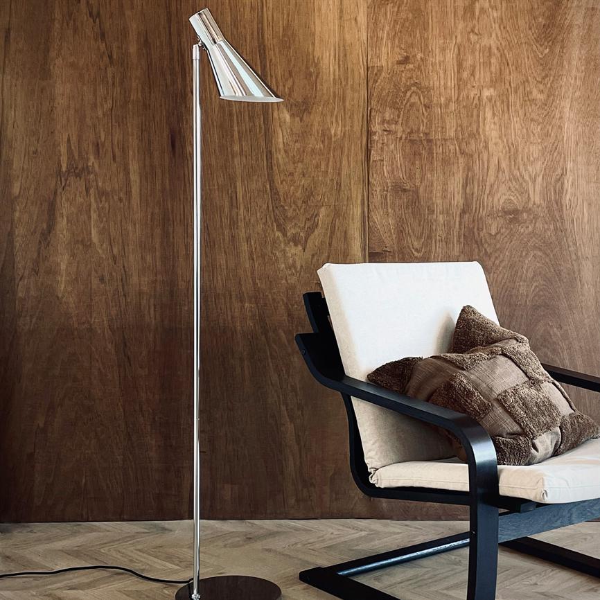 A chrome floor lamp stands next to an upholstered armchair with a cream cover and brown throw pillows, in front of a warm dark wooden wall and light patterned wooden floor.