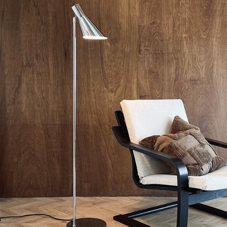 A floor lamp is lit next to an upholstered armchair with a beige cushion and brown cushions, in front of a warm wood-clad wall and light herringbone parquet floor.