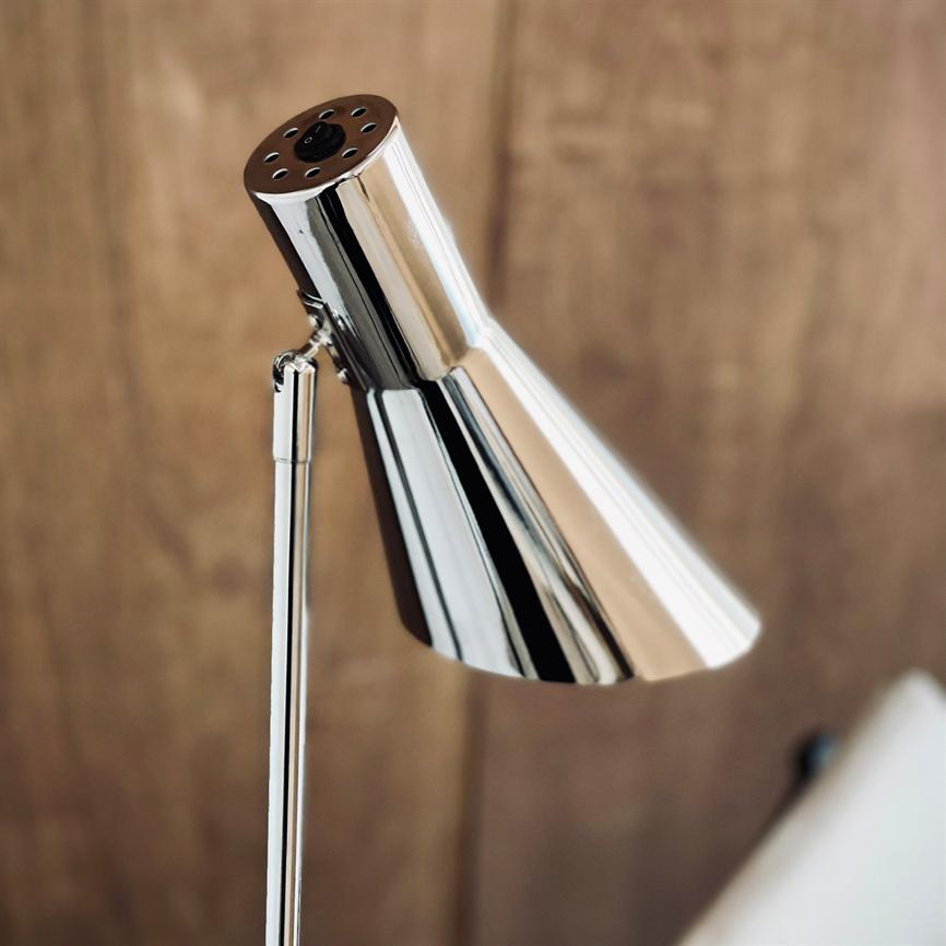 A chrome-colored lamp head tilts and points downwards; a slender metal rod supports it, in front of a blurred wood-clad background with a partially visible cushion in the lower right corner.