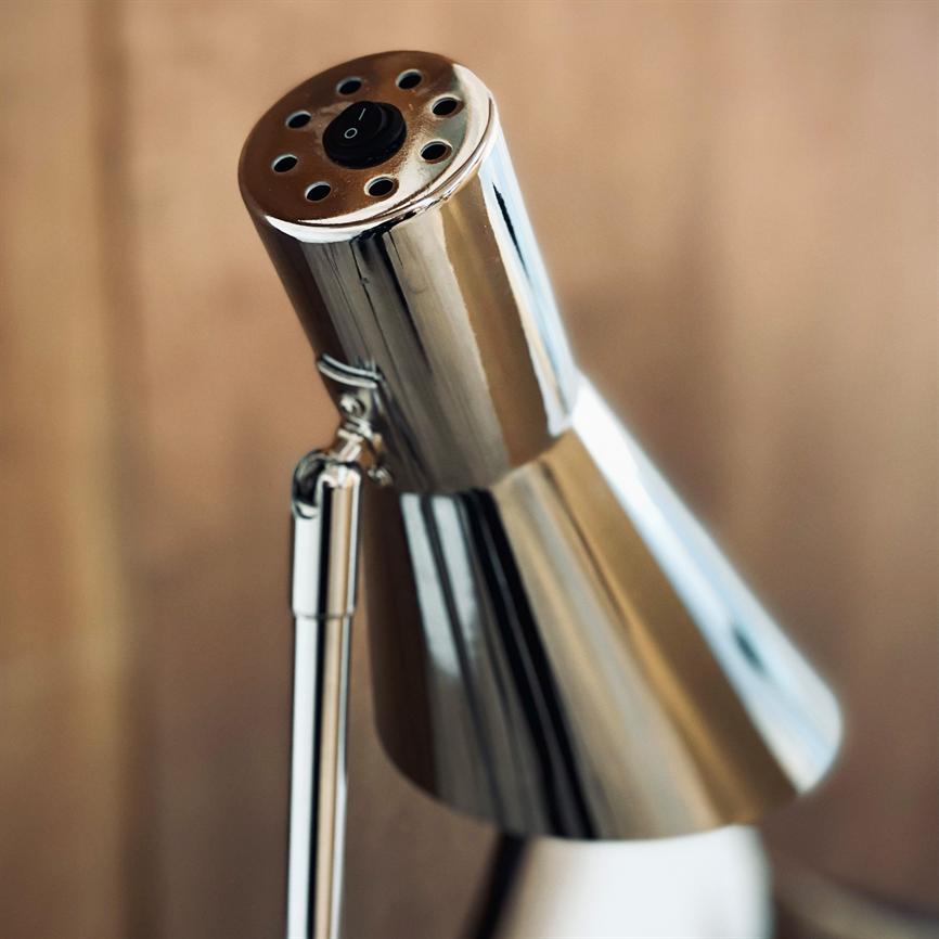 A chrome table lamp — the lamp head is angled forward and reflects light, with ventilation holes and a black switch marked "I" — in front of a blurred, light wood background in close-up.