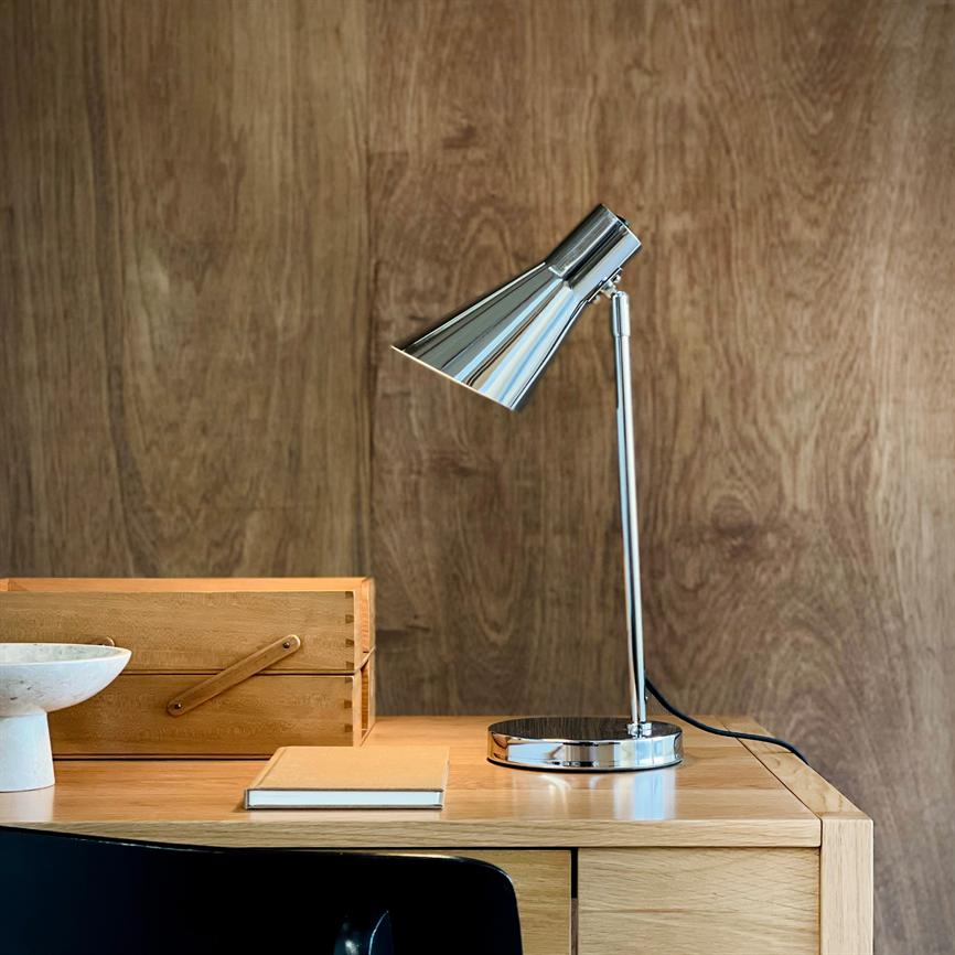 Object: chrome desk lamp. Action: stands angled downwards above the work surface. Context: wooden desk with notebook, wooden box and bowl in front of a warm, wood-clad wall.